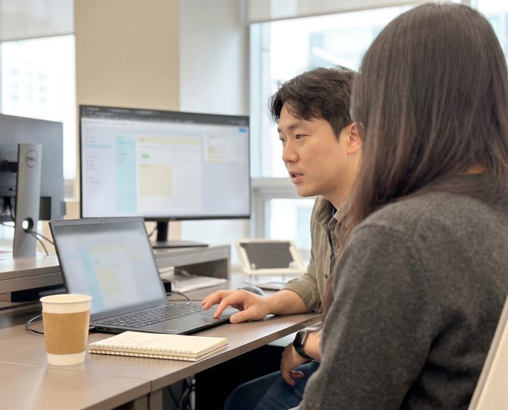 한 사람이 노트북 화면을 가리키며 옆에 앉은 담당자에게 설명합니다.